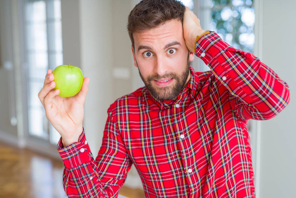 Hombre guapo comiendo manzana verde fresca y saludable estresado con la mano en la cabeza, conmocionado con vergüenza y cara sorpresa, enojado y frustrado. Miedo y malestar por error
. - Foto, Imagen
