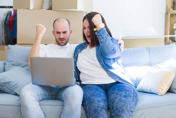 Casal jovem sentado no sofá arround caixas de papelão se movendo para uma nova casa usando laptop irritado e frustrado gritando com raiva, louco e gritando com a mão levantada, conceito de raiva
 - Foto, Imagem