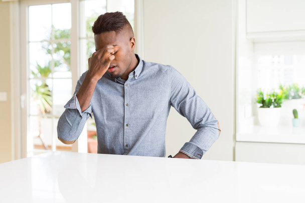 Handsome african american man on white table tired rubbing nose and eyes feeling fatigue and headache. Stress and frustration concept. - Photo, Image