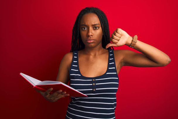 junge afrikanisch-amerikanische Studentin liest Buch vor isoliertem roten Hintergrund mit wütendem Gesicht, negatives Zeichen für Abneigung mit gesenktem Daumen, Ablehnungskonzept - Foto, Bild