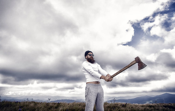 Hipster with beard on strict face holds axe, cloudy sky background. Man with beard holds axe while stand on top of mountain. Hiking concept. Hipster looks brutal and stylish with axe and sunglasses - Zdjęcie, obraz