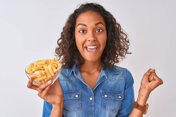 Young brazilian woman holding bowl with macaroni pasta over isolated white background screaming proud and celebrating victory and success very excited, cheering emotion - Foto, Imagen
