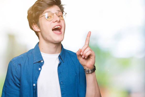 Young handsome man wearing glasses over isolated background pointing finger up with successful idea. Exited and happy. Number one. - Photo, Image