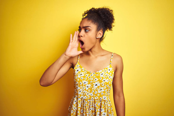 Afro-Amerikaanse vrouw het dragen van casual bloemen jurk staande over geïsoleerde gele achtergrond schreeuwen en schreeuwen luid aan de kant met de hand op de mond. Communicatie concept. - Foto, afbeelding