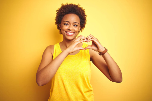 愛に微笑む孤立した黄色の背景の上に夏のTシャツを着たボーイトゥルアフリカ系アメリカ人女性が手でハートのシンボル形状をしています。ロマンチックなコンセプト. - 写真・画像