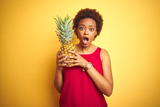 African American kobieta trzyma tropikalny ananas na żółtym białym tle przestraszony w szoku z niespodzianką twarz, boi się i podekscytowany wyrazem strachu - Zdjęcie, obraz