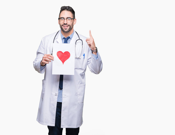 Красивый молодой врач держит бумагу с красным сердцем на изолированном фоне, удивленный идеей или вопросом, указывая пальцем со счастливым лицом, номер один
 - Фото, изображение