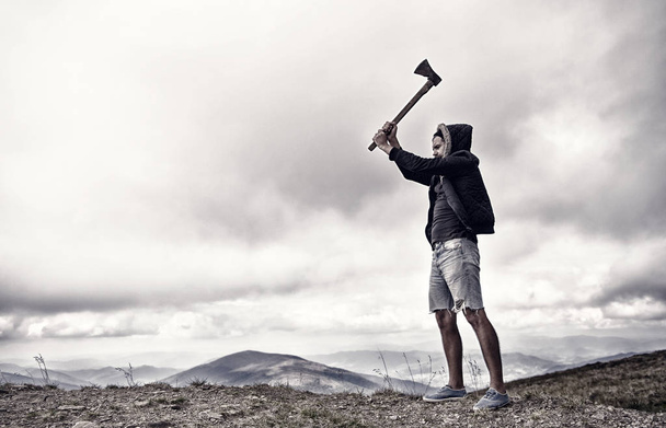 Hipster with beard holds axe while stand on top of mountain, sky on background, copy space. Lumberjack brutal and bearded raising axe. Man in hat and jacket conquered top. Brutal lumberjack concept - Foto, imagen