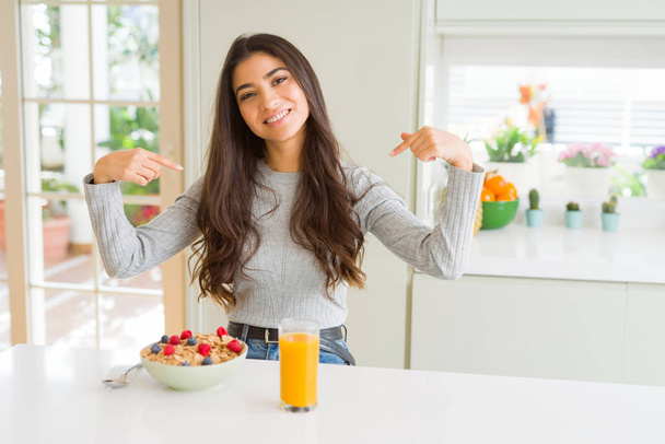 Mujer joven desayunando sano por la mañana mirando confiado con sonrisa en la cara, señalándose con los dedos orgullosos y felices
. - Foto, Imagen