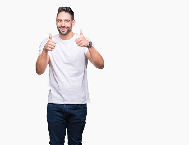 Mladý muž nosí ležérní bílé tričko přes znamení úspěchu izolované pozadí dělá pozitivní gesto rukou, palec se usmívá a šťastný. Při pohledu na fotoaparát s veselým výrazem, vítěz gesto. - Fotografie, Obrázek