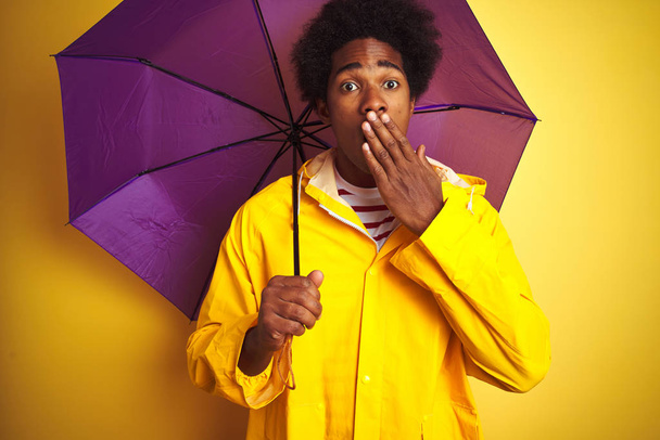 Afro amerikan adam yağmur ceket ve şemsiye giyen izole sarı arka plan kapağı ağız üzerinde duran el hata için utanç ile şok, korku ifadesi, sessizlik, gizli kavram korkmuş - Fotoğraf, Görsel