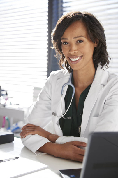 Portrait Of Smiling Female Doctor Wearing White Coat With Stethoscope Sitting Behind Desk In Office - 写真・画像