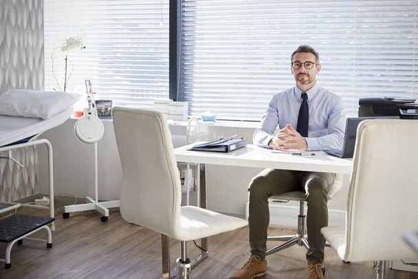 Portrait Of Mature Male Doctor Sitting Behind Desk In Office - Photo, image
