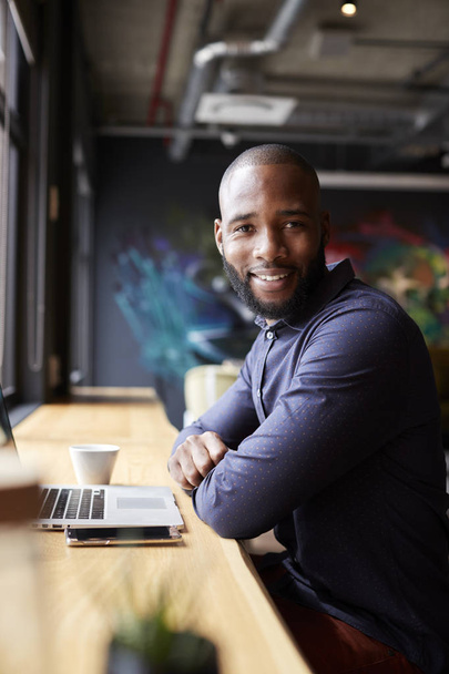 Черный мужчина креативно сидит у окна в кафе с ноутбуком, поворачивается и улыбается камере, вертикально
 - Фото, изображение