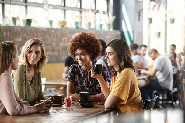 Four Young Female Friends Meeting Sit At Table In Coffee Shop And Talk - 写真・画像
