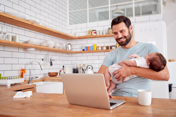 Multi-Tasking Father Holding Sleeping Baby Son And Working On Laptop Computer In Kitchen - Foto, Imagem