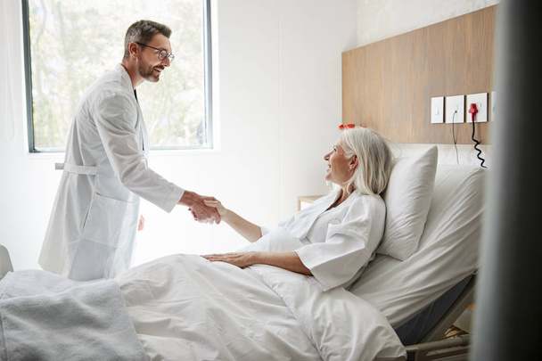Surgeon Visiting And Shaking Hands With Mature Female Patient In Hospital Bed - 写真・画像