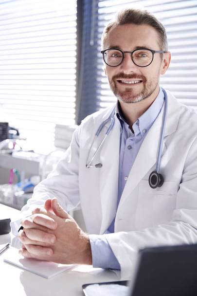 Porträt eines lächelnden Arztes in weißem Mantel mit Stethoskop, der hinter Schreibtisch im Büro sitzt - Foto, Bild