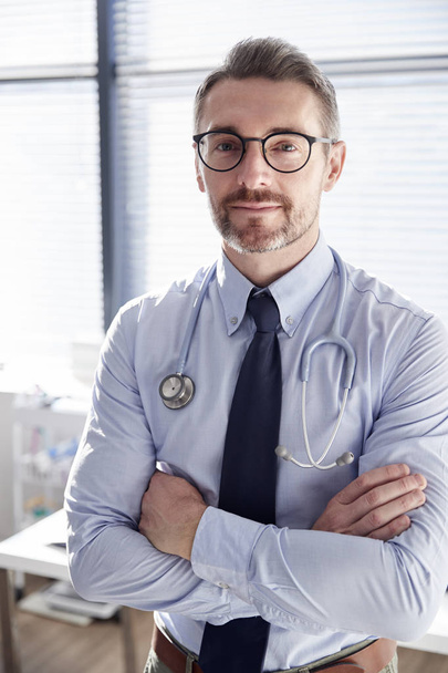 Portrait Of Smiling Mature Male Doctor With Stethoscope Standing By Desk In Office - Foto, Imagem