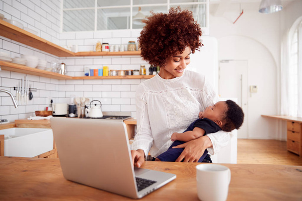 Multi-Tasking Mother Holding Sleeping Baby Son And Working On Laptop Computer In Kitchen - Foto, Imagem