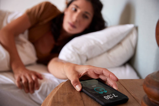 Mid adult woman waking up in bed, reaching out to smartphone on the bedside table in the foreground - Photo, Image