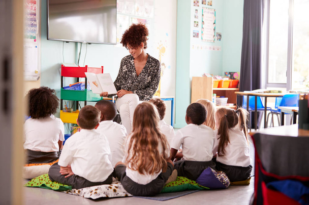 Female Teacher Reading Story To Group Of Elementary Pupils Wearing Uniform In School Classroom - Foto, imagen