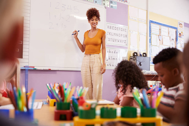 Female Teacher Standing At Whiteboard Teaching Maths Lesson To Elementary Pupils In School Classroom - Foto, Imagem