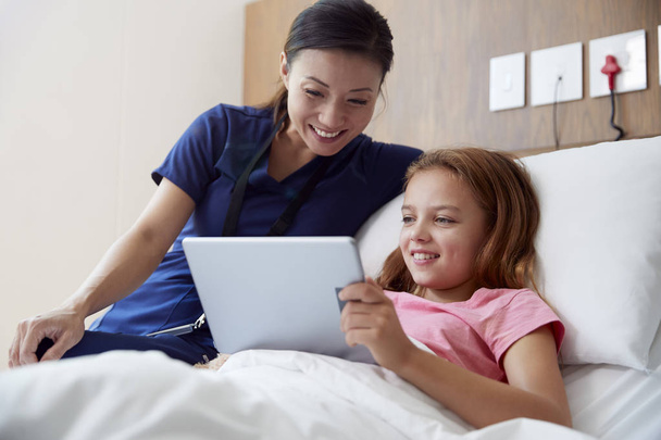 Female Nurse With Girl Lying In Hospital Bed Looking At Digital Tablet Together - Φωτογραφία, εικόνα