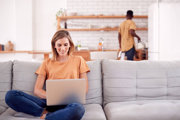 Woman Relaxing Sitting On Sofa At Home Using Laptop Computer With Man In Kitchen Behind - Foto, Bild