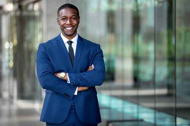 オフィスの外の職場、カラフルな反射ガラスの建物、コピースペースを横切った腕で誇りに立つ笑顔のアフリカ系アメリカ人ビジネスマンCEO - 写真・画像