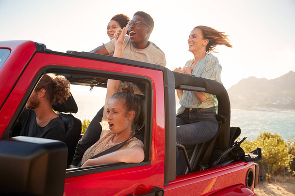 Five young adult friends on a road trip driving in an open top jeep by the sea - Фото, изображение