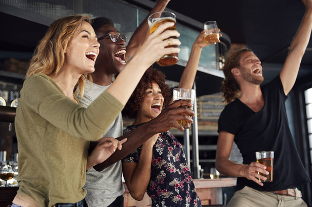 Group Of Male And Female Friends Celebrating Whilst Watching Game On Screen In Sports Bar - Foto, afbeelding