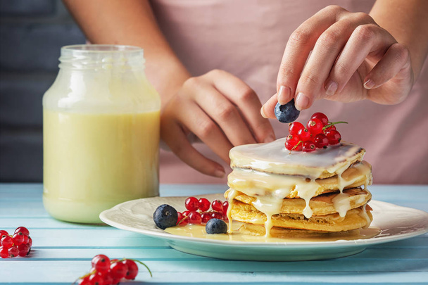 A girl in a pink apron decorates pancakes with blueberries and red currants. Tasty pancakes with condensed milk. Close-up photograph of fresh natural dessert - Photo, Image