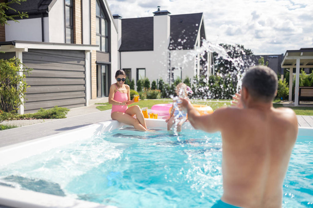 Father splashing water on his daughter and wife in the swimming pool - Photo, Image
