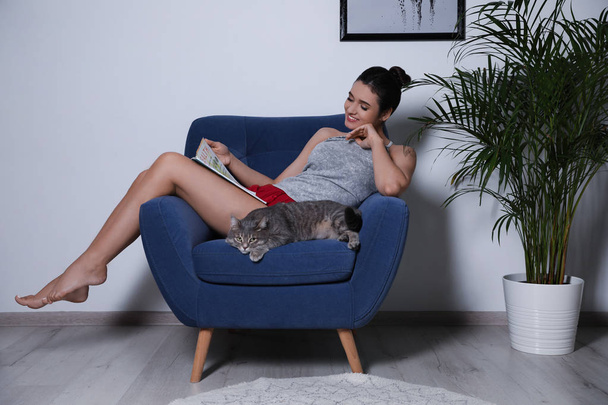 Young woman with cat and magazine on armchair at home. Cute pet - Foto, afbeelding