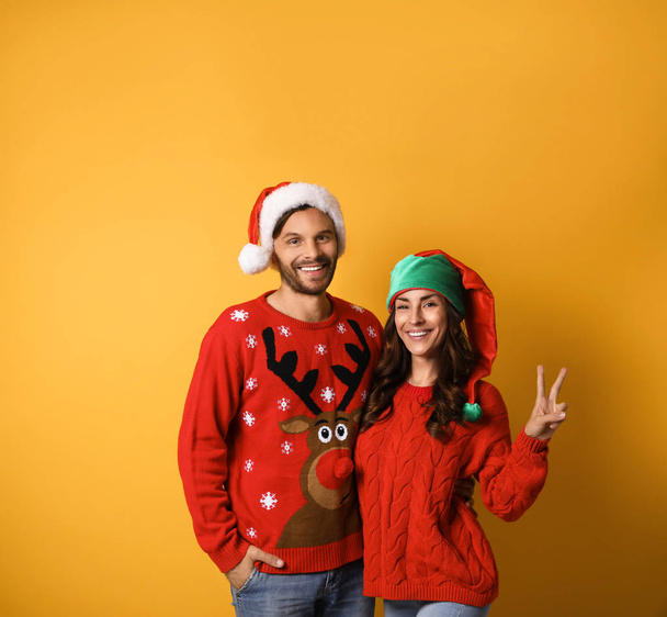 Young couple in Christmas sweaters and hats on yellow background. Space for text - Zdjęcie, obraz