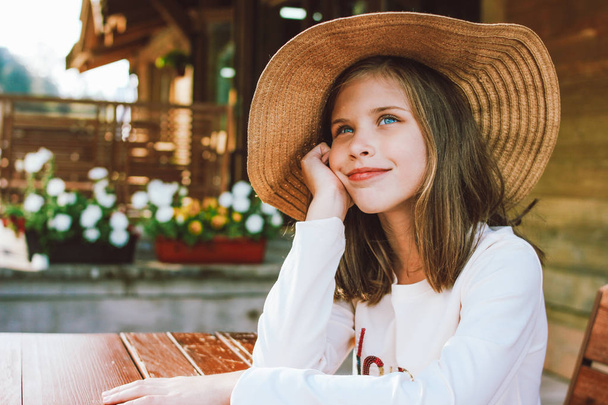 Attractive preteen girl in straw hat with blue eyes sitting and dreaming at table in backyard of wooden cottage - Photo, Image