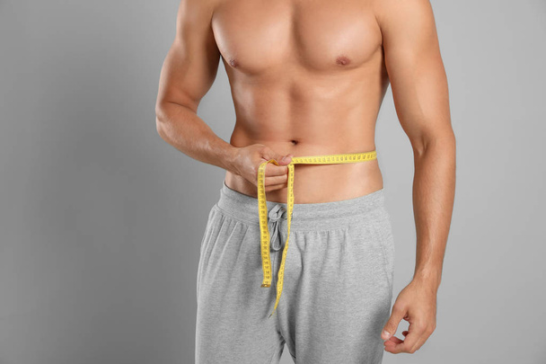 Young man with slim body using measuring tape on grey background, closeup view - Foto, afbeelding