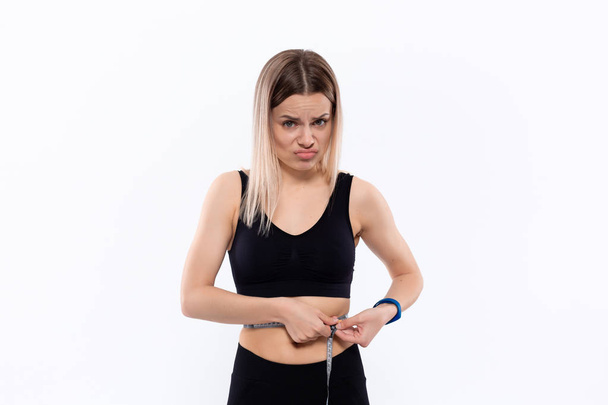 Jonge blonde vrouw in een zwarte sportkleding met slimme horloges is boos controleren van de taille na de training met de meetlint staande over witte achtergrond. - Foto, afbeelding