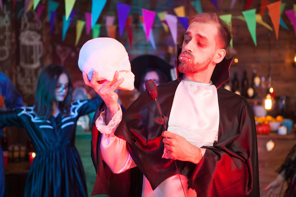 attraktiver junger Mann, der wie Dracula verkleidet ist und eine schwarze Rose in der Hand hält und seinen menschlichen Schädel bei der Halloween-Feier betrachtet - Foto, Bild