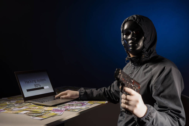 Ein anonymer Hacker-Programmierer benutzt einen Laptop mit einer Pistole in der Hand, um das System im Dunkeln zu hacken. Entstehung und Infektion von bösartigen Viren. - Foto, Bild