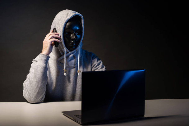 Anonieme hacker in Mask programmeur maakt gebruik van een laptop en praten op de telefoon om het systeem te hacken in het donker. - Foto, afbeelding