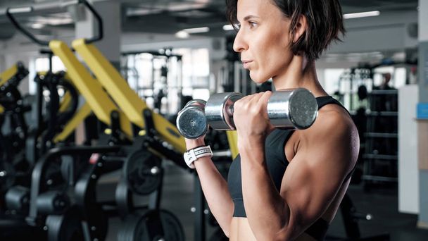 Sporty mature woman is making biceps exercise with dumbbells in gym. - Photo, Image