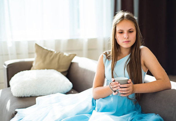 Porträt eines jungen Teenagers brünettes Mädchen mit langen Haaren, das zu Hause auf einem Sofa mit Tasse sitzt - Foto, Bild