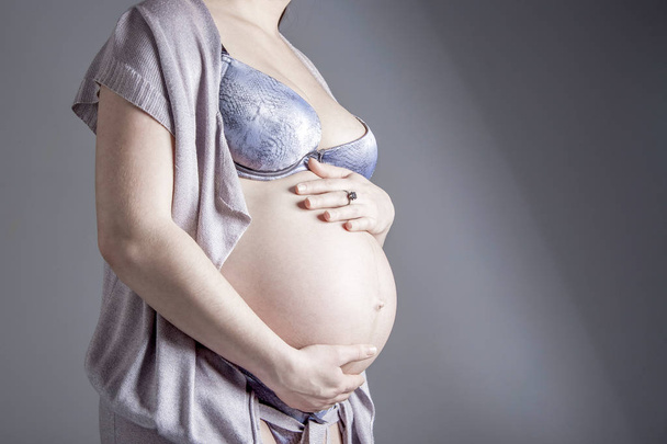 Pregnant woman on gray background in studio holding hands on her - Φωτογραφία, εικόνα