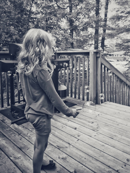 Чорно-білий портрет блондинки дошкільного віку, яка вискакує мильні бульбашки на дерев'яній палубі на подвір'ї в котеджі. Літо весело. Щасливий спосіб життя в дитинстві. Вид ззаду
 - Фото, зображення