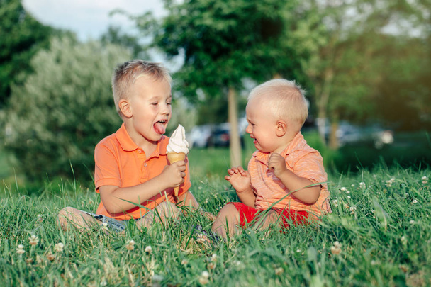 Δύο Καυκάσιοι αστεία παιδιά αγόρια κάθονται μαζί τρώγοντας ένα παγωτό. Νήπιο νεότερο μωρό κλάμα και μεγαλύτερος αδελφός πειράγματα του. Αγάπη ζήλια ζηλιάρης αδέλφια φιλία. - Φωτογραφία, εικόνα