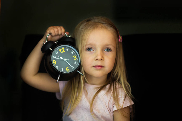 Back to school concept. A schoolgirl with an alarm clock in hand is shocked. A small girl is surprised and looking up with a black clock with a hand holding her head on a black background. - Foto, afbeelding