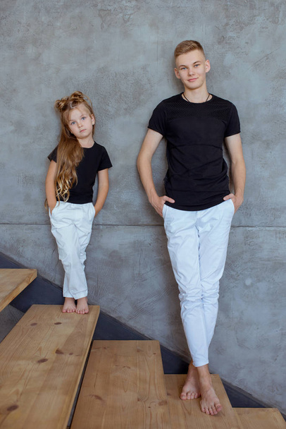 stijlvolle Kaukasische broers en zussen (broer en zus) in blak t-shirts en witte broek bij de grijze muur in loft interieur. Familie, mode, familie, relatie en kinderconcept. - Foto, afbeelding