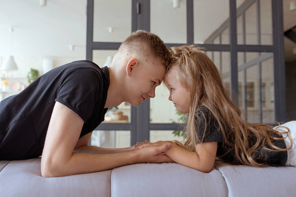 caucasian siblings - teenager boy brother and little girl sister laying holding hands on a couch (sofa) in modern loft interior - Foto, Imagem